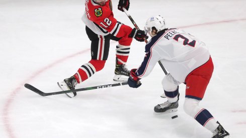 Feb 13, 2021; Chicago, Illinois, USA; Chicago Blackhawks left wing Alex DeBrincat (12) shoots between the legs of Columbus Blue Jackets defenseman Andrew Peeke (2) during the second period at the United Center.